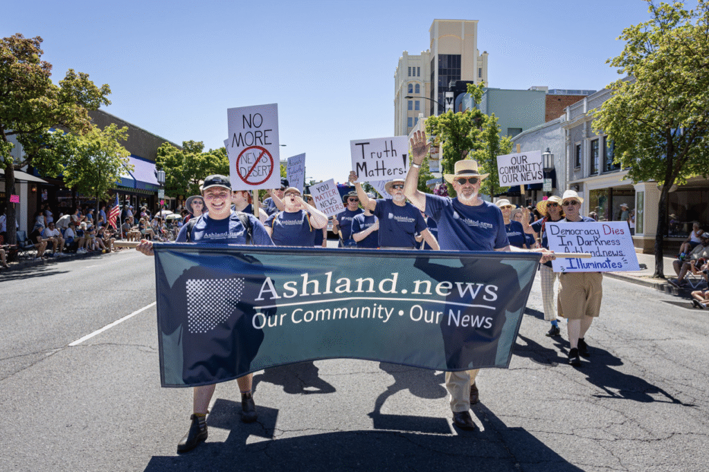 "Ashland.news team members and supporters marching in a downtown parade on a sunny day. The group carries a large banner reading 'Ashland.news: Our Community, Our News' and holds signs supporting independent journalism with slogans like 'Truth Matters,' 'No More News Desert,' and 'Democracy Dies in Darkness.'"